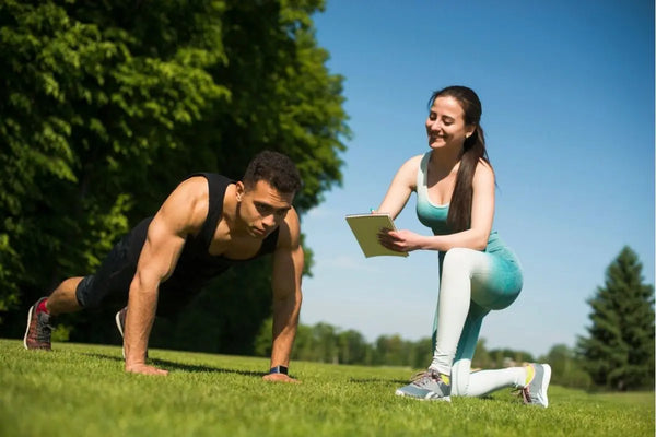 seorang pria sedang push up di outdoor dengan wanita di sisinya memegang buku