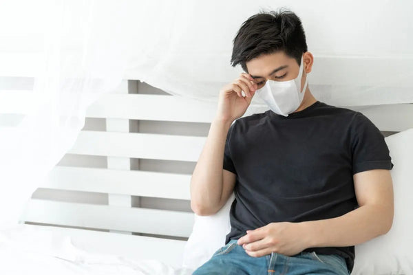 orang yang duduk di tempat tidur sambil memegang kepala dan memakai masker