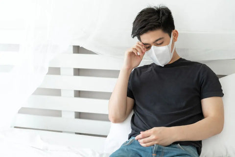 orang yang duduk di tempat tidur sambil memegang kepala dan memakai masker